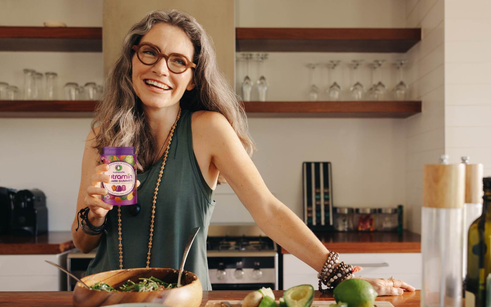 a smiling woman leaning on a counter is holding an open bottle of nutramin vegan multivitamins