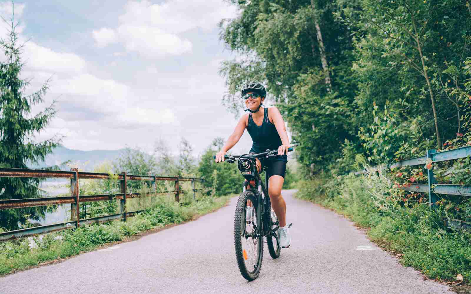 A smiling woman is on a bike in the countryside, surrounded by lush greenery.  She's wearing a helmet, sunglasses, and a sports shirt/shorts that are all black. She's smiling.