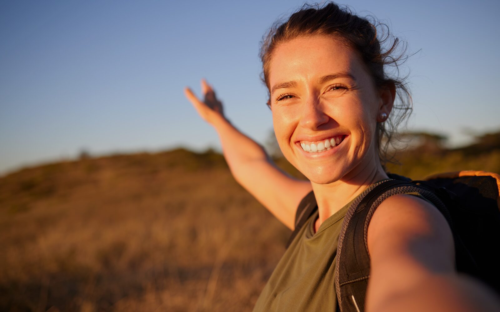 A woman is smiling in the evening sun - gesturing towards an open field. She's wearing a backpack and looks healthy/fit.