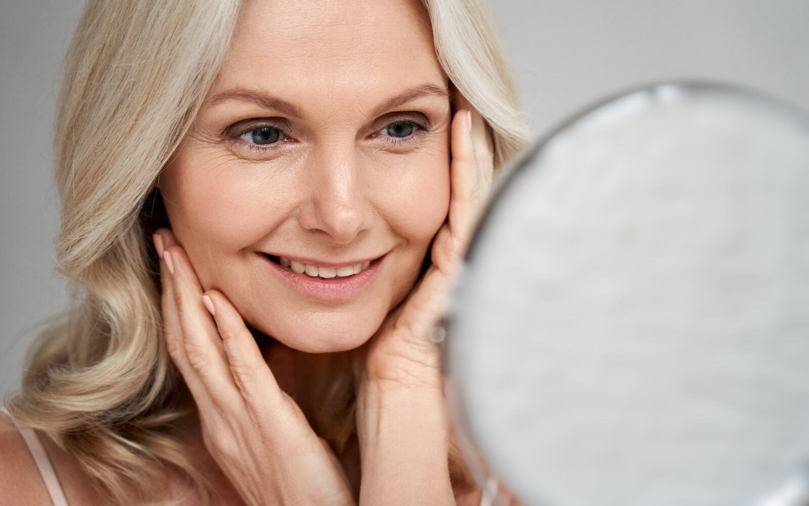 Happy 50s middle aged woman model touching face with beautiful skin looking in mirror.