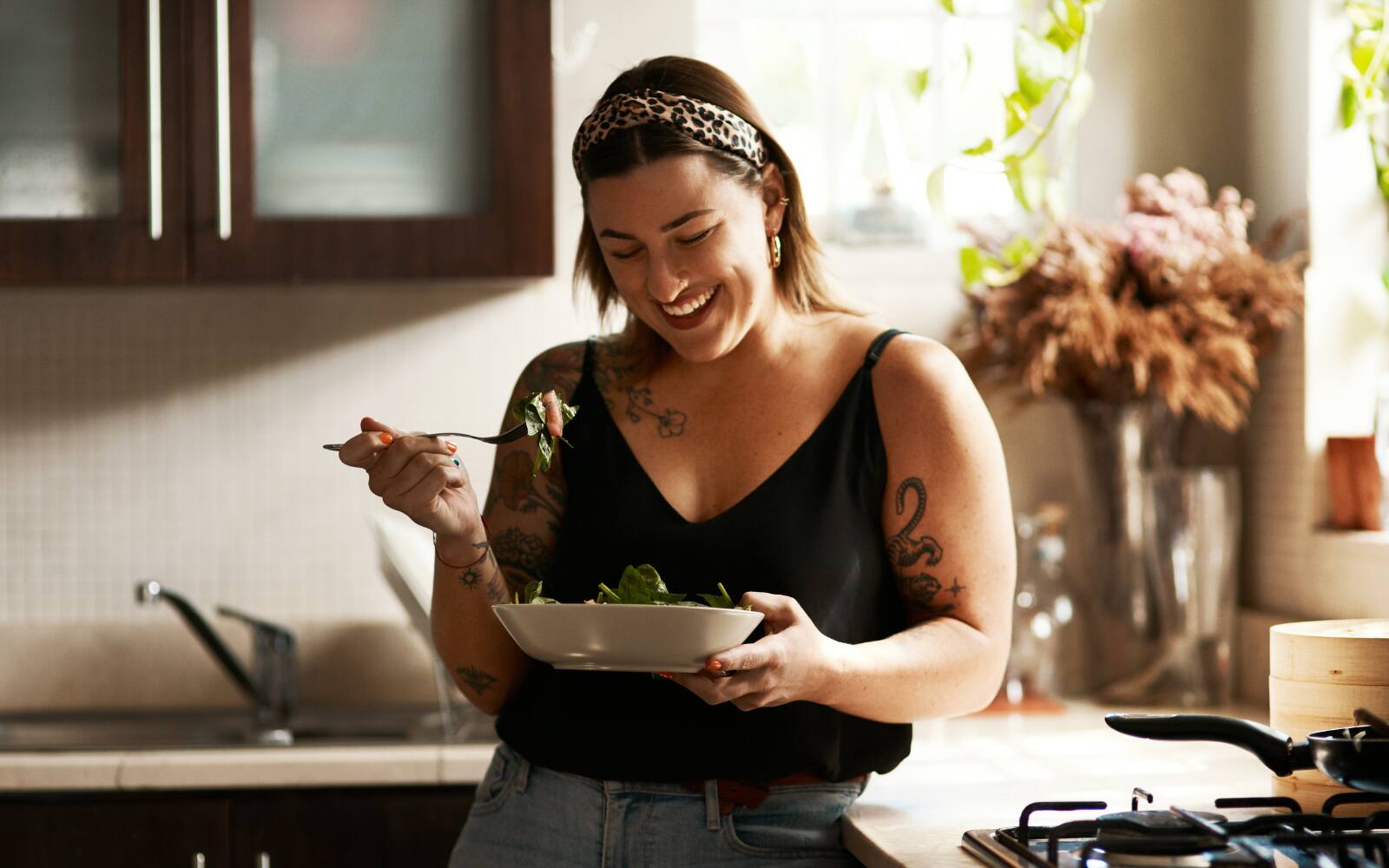 A woman is smiling in her kitchen holding a bowl of healthy-looking food. The image gives happy and healthy vibes.