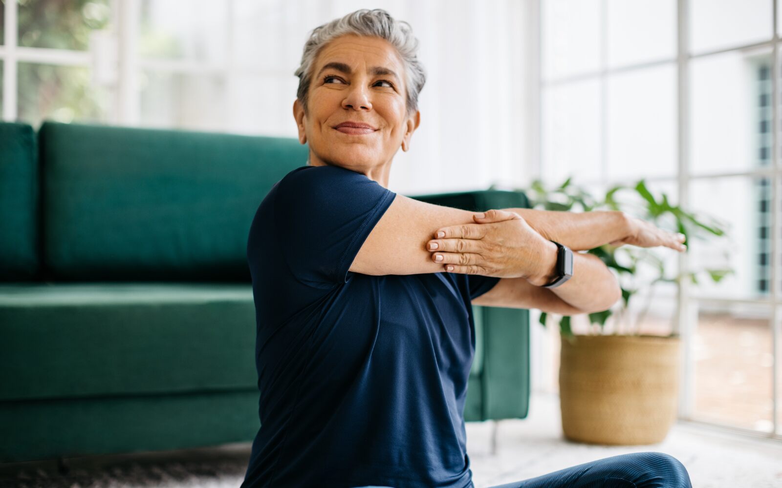 A short-haired woman is stretching while sitting on her living room floor.  She looks healthy and content.