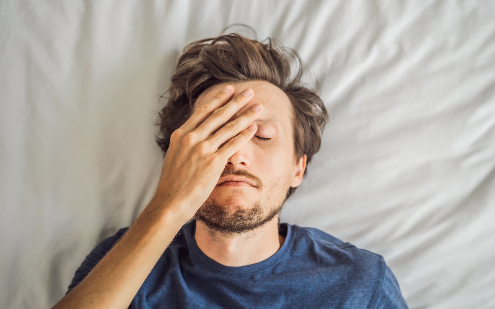 a man is laying on a white bed, with his face over his hand, there are no words on the image - he looks tired
