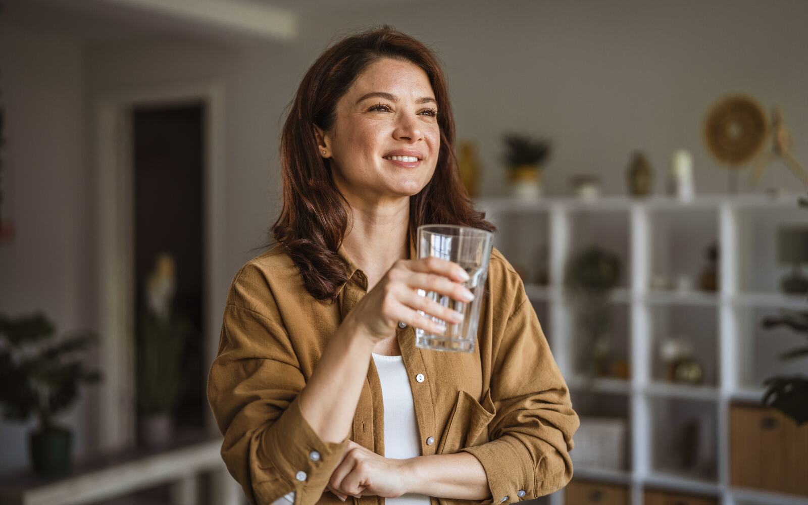 A woman is standing in her brightly-lit room, with a glass of water, she's smiling  and has healthy, clear skin
