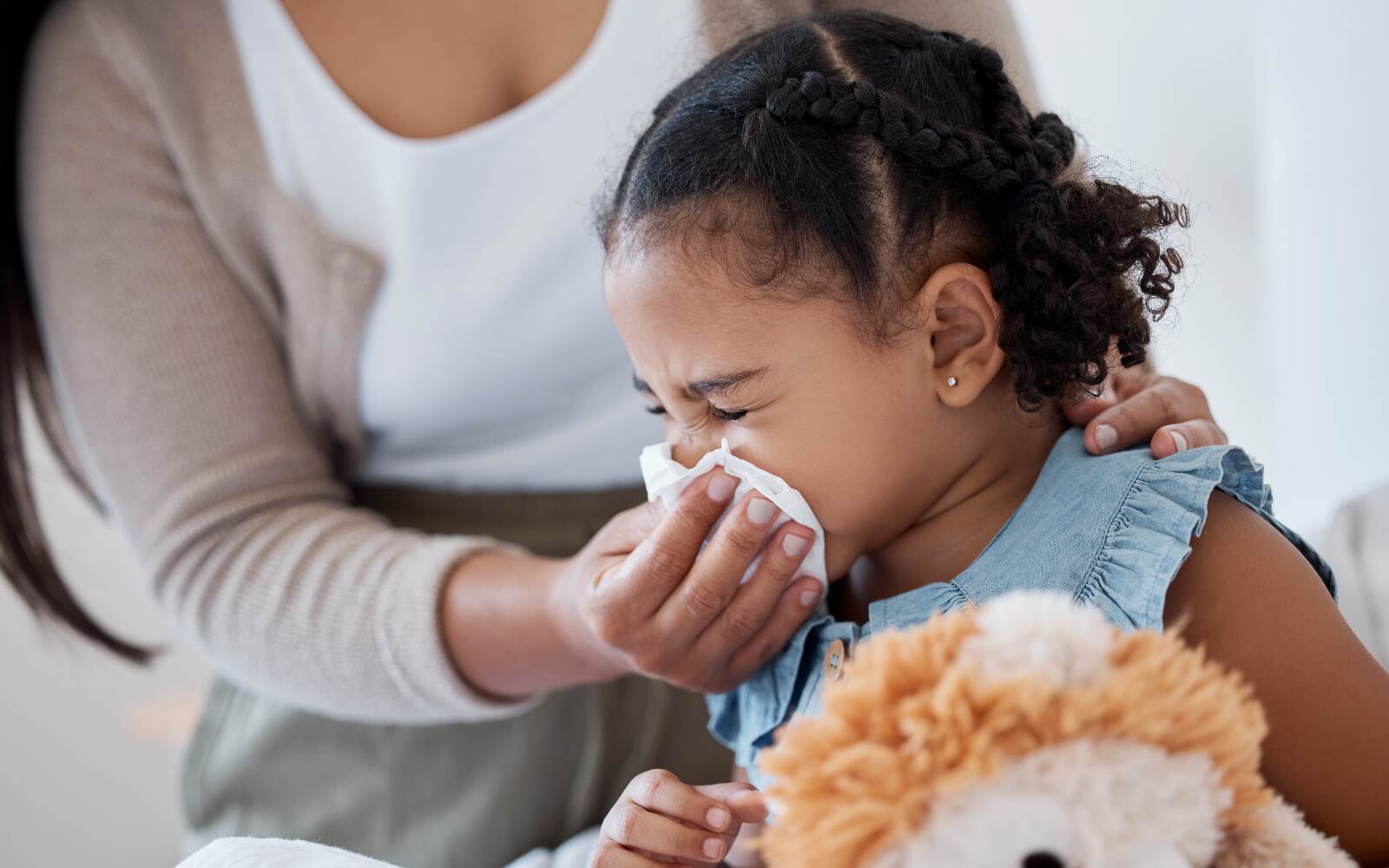 A young girl blows her nose into a tissue while an adult comforts her with a hand on her shoulder. A stuffed animal sits beside her.