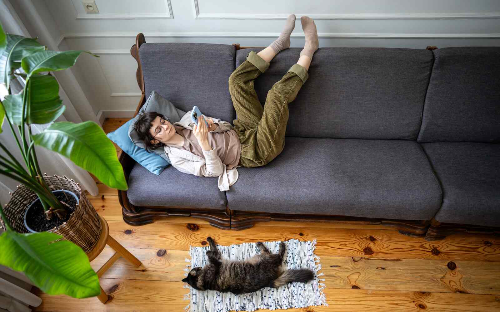 A woman is lounging on a couch on a sunny afternoon, with a cat snoozing beside her on the floor. The vibe is chill.