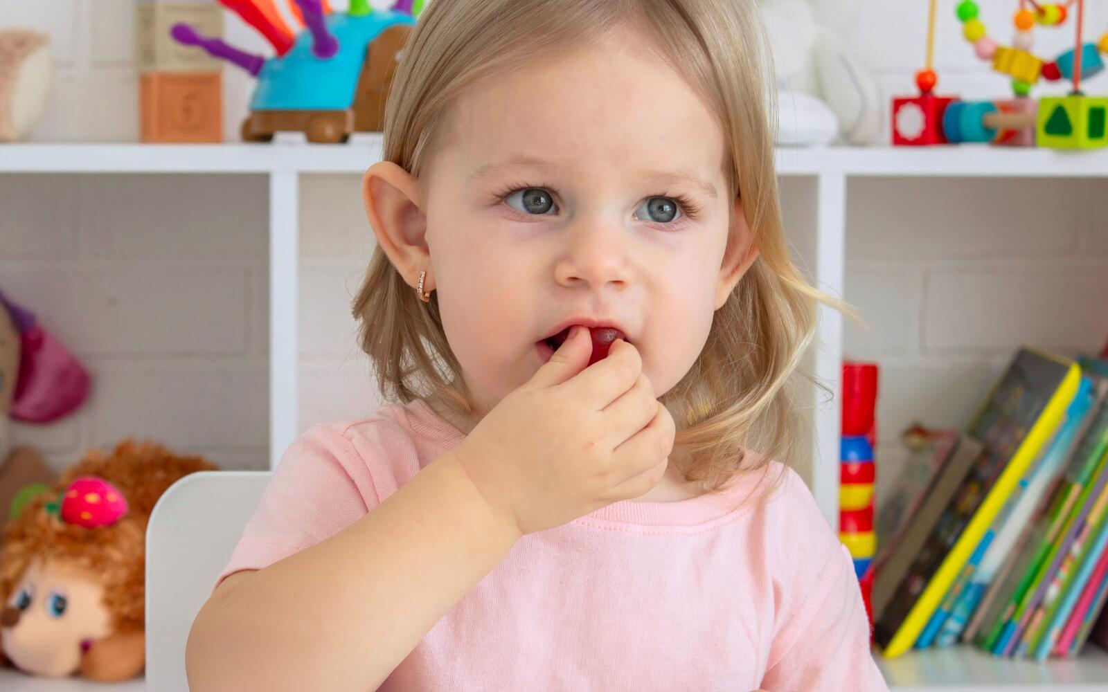 a young girl with blonde hair and blue eyes is biting into a yummy-looking red vitamin gummy. 