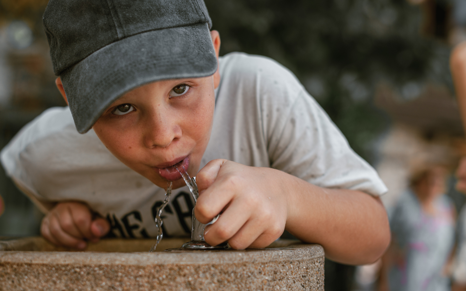 Boy in a gray cap and white t-shirt drinking water from a public fountain on a sunny summer day. A candid and refreshing outdoor moment.