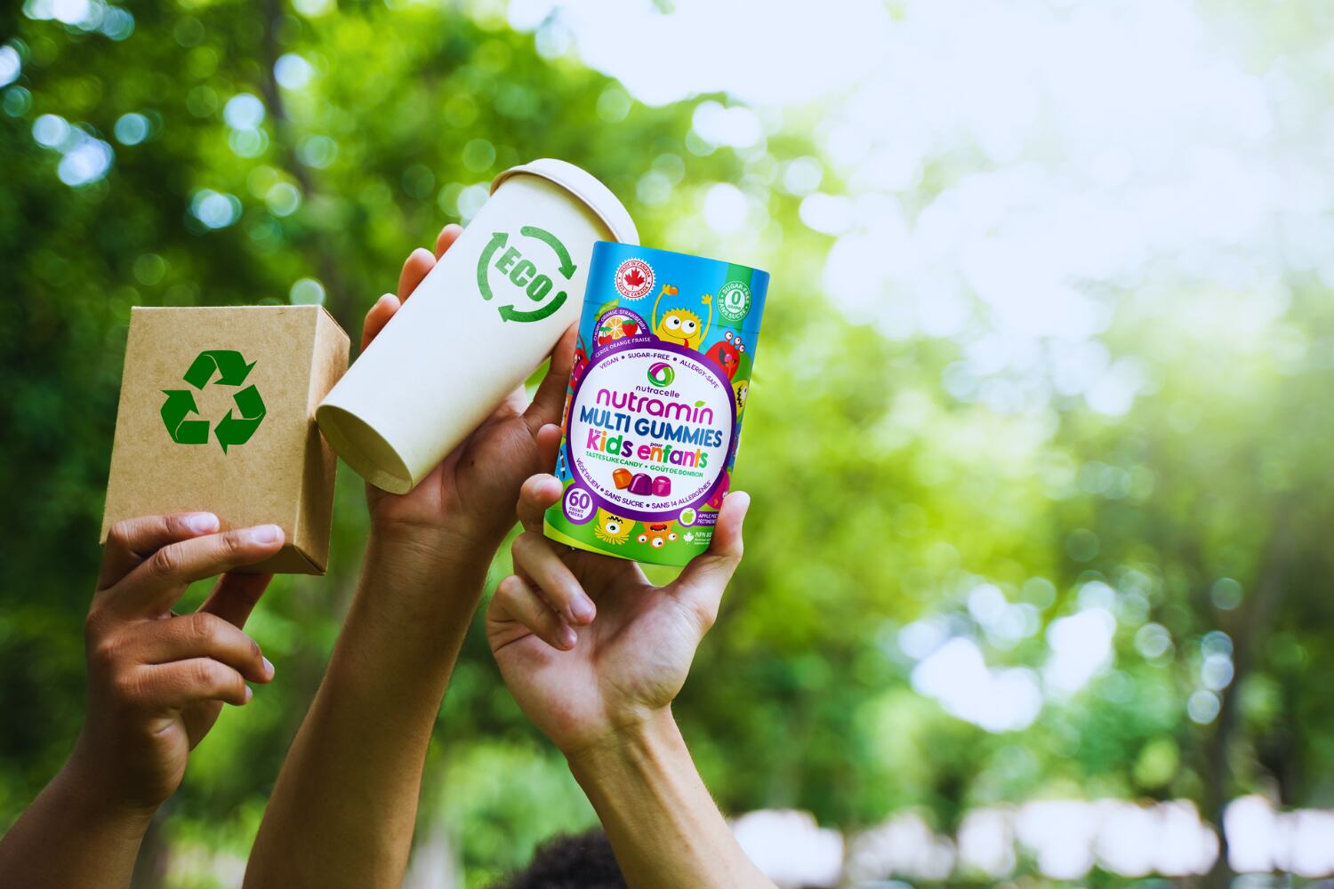 Three hands are holding eco-friendly products: a recycleable coffee cup, a bottle of kids Nutramin vegan multivitaamins and a box that has the reduce reuse recycle logo