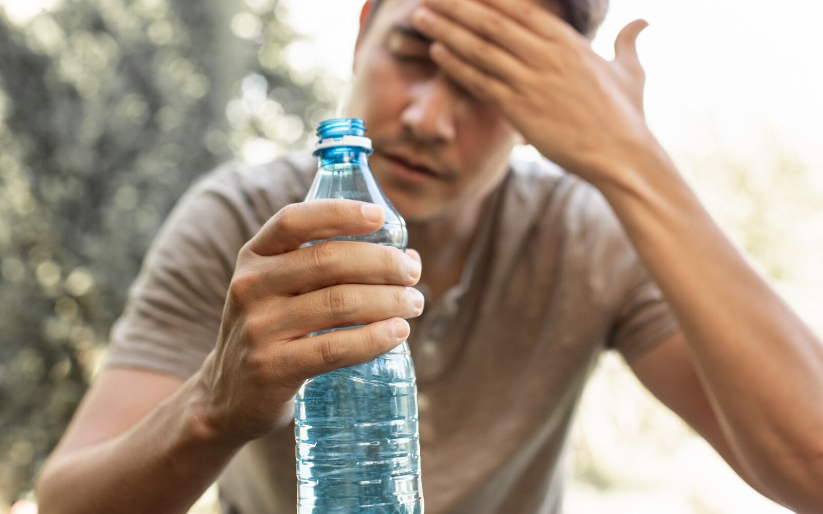 a man is outside, holding a bottle of water with his other hand on his forehead.  He looks tired and in need of a drink.