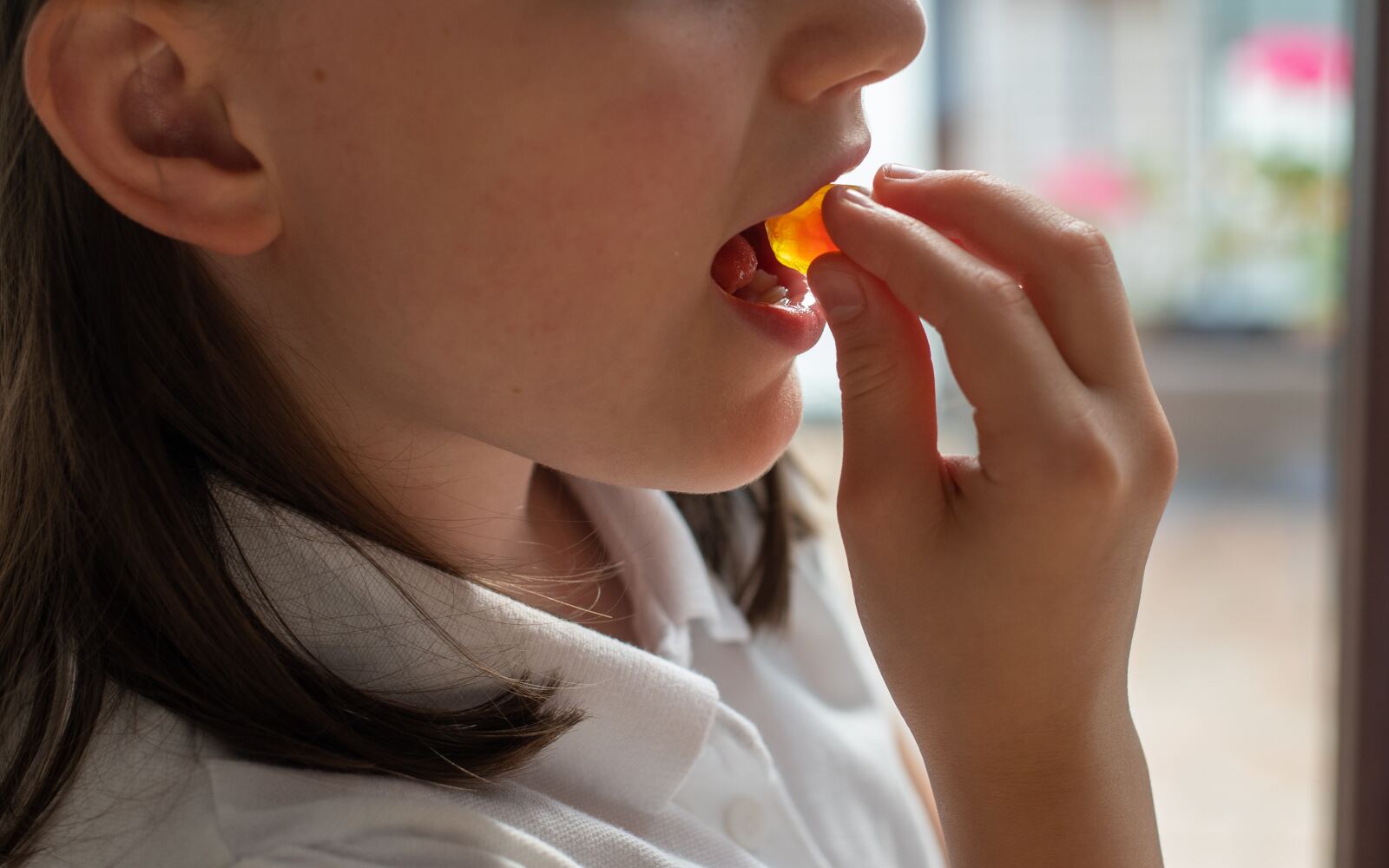 a child is putting an orange vitamin gummy in their mouth, the photo is cropped closely. you can only see their hand, the gummy, and from their nose down