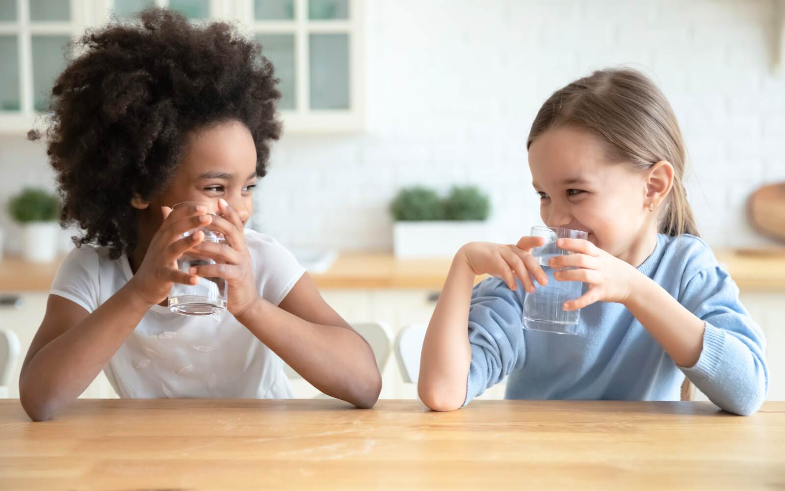 two kids are smiling at each other while holding clear glasses with water in front of their faces.  They look like they were mid-drink when they started giggling together.