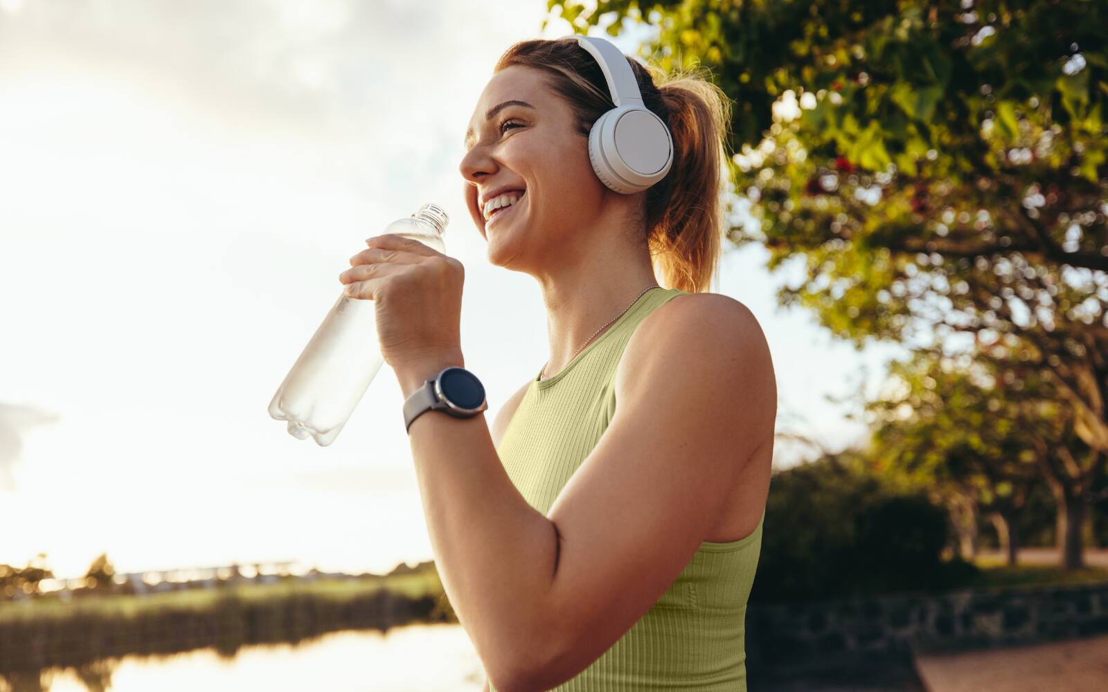 a woman is smiling, holding a bottle of water while wearing an active wear top and headphones, the background looks like a lake nearby, she's possibly out for a fitness run.