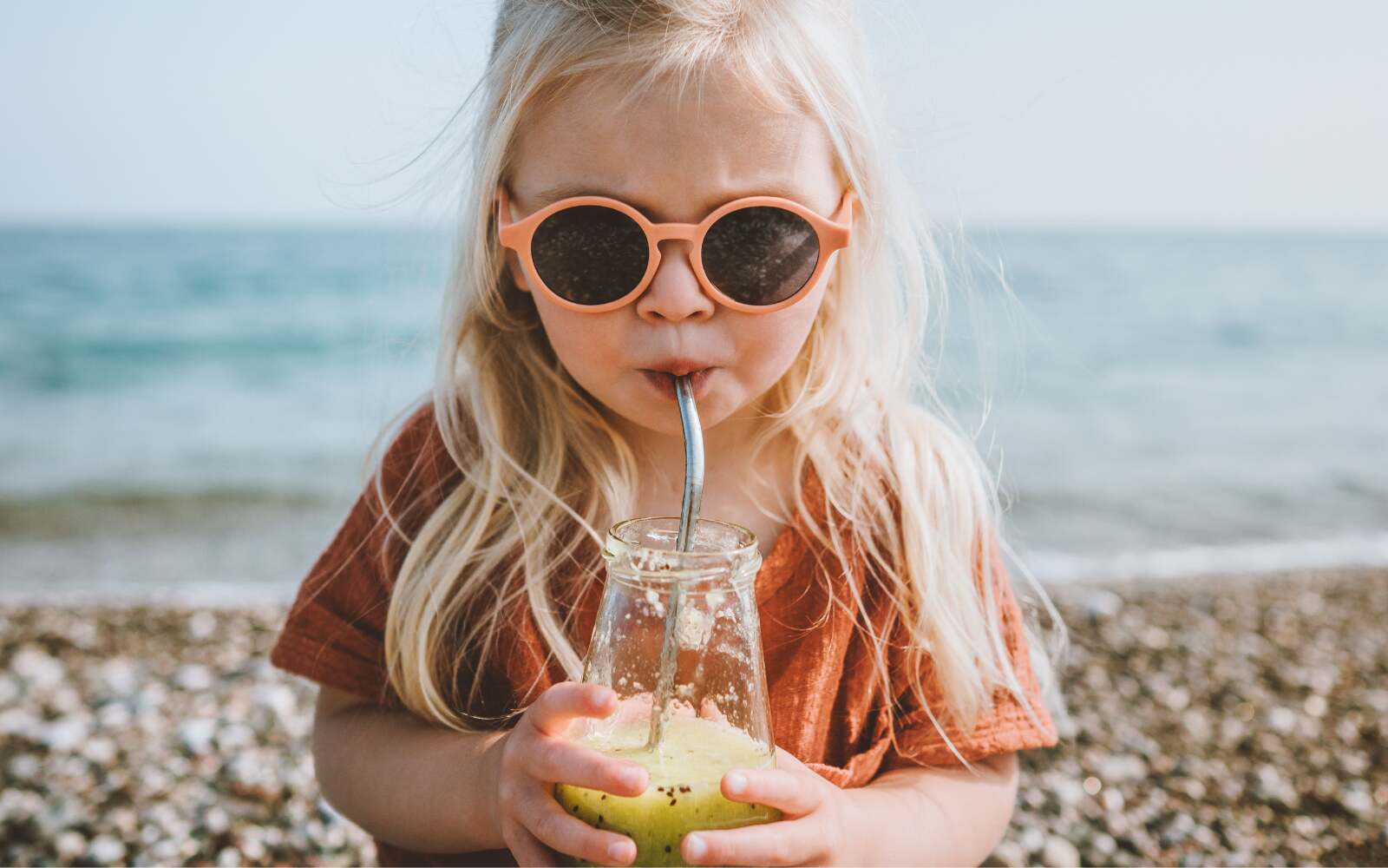 a child with sunglasses and long blonde hair is drinking a  healthy-looking juice