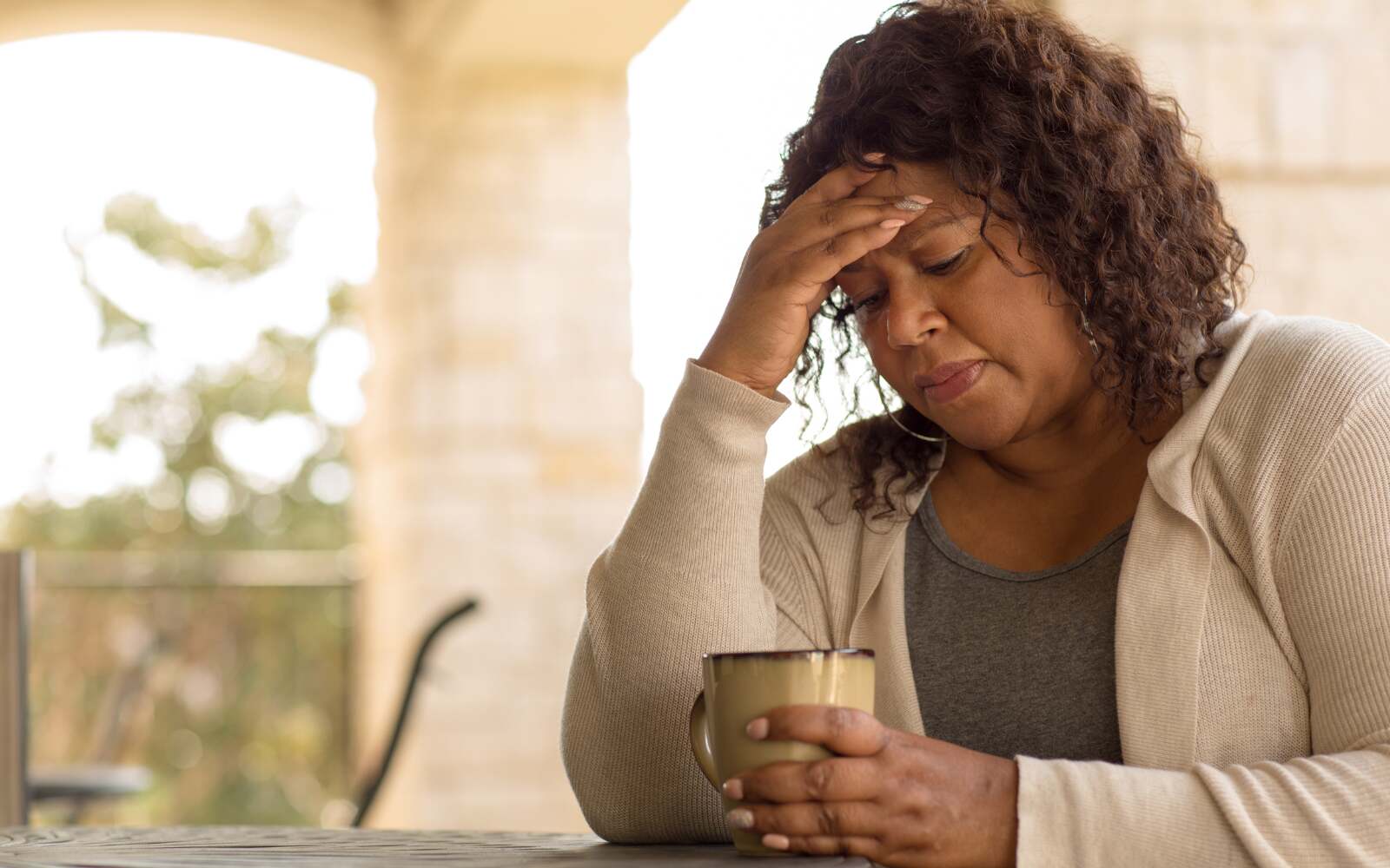 Middle-aged woman sitting at a table holding a cup of coffee and touching her forehead, appearing tired or stressed — representing early signs of perimenopause.