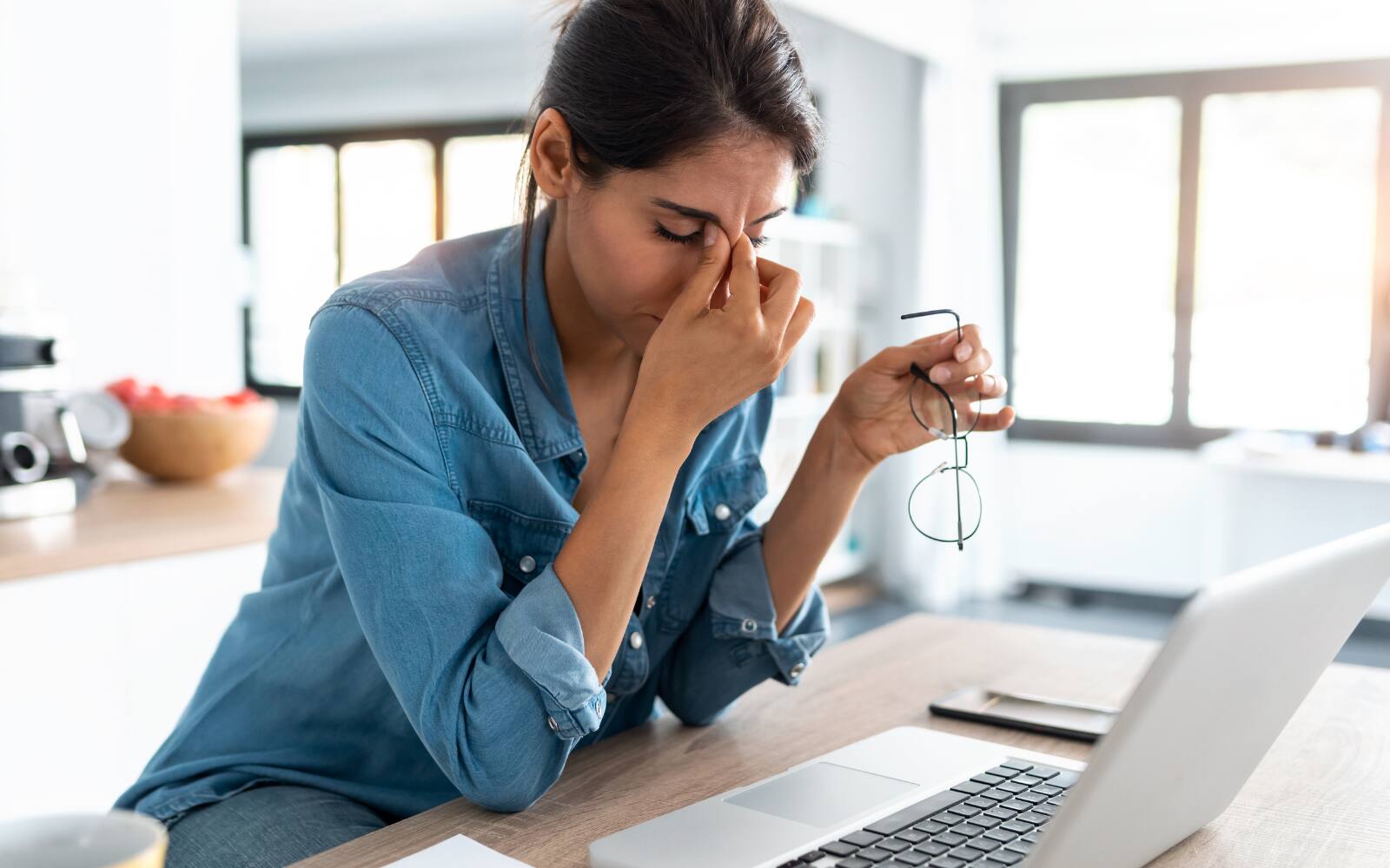 A woman is pinching the bridge of her nose with her right hand, while holding her glasses in the other. She's sitting in front of a laptop looking visibly stressed.