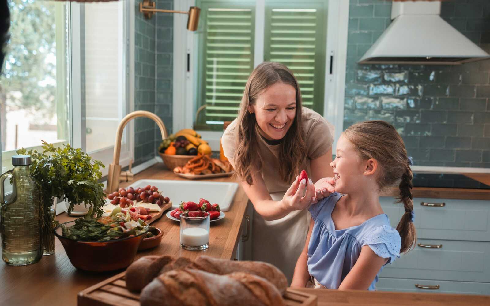 a mom and daughter are in their kitchen, smiling next to a counter full of freshly-washed strawberries.  The mom is feeding a strawberry to the smiling girl.