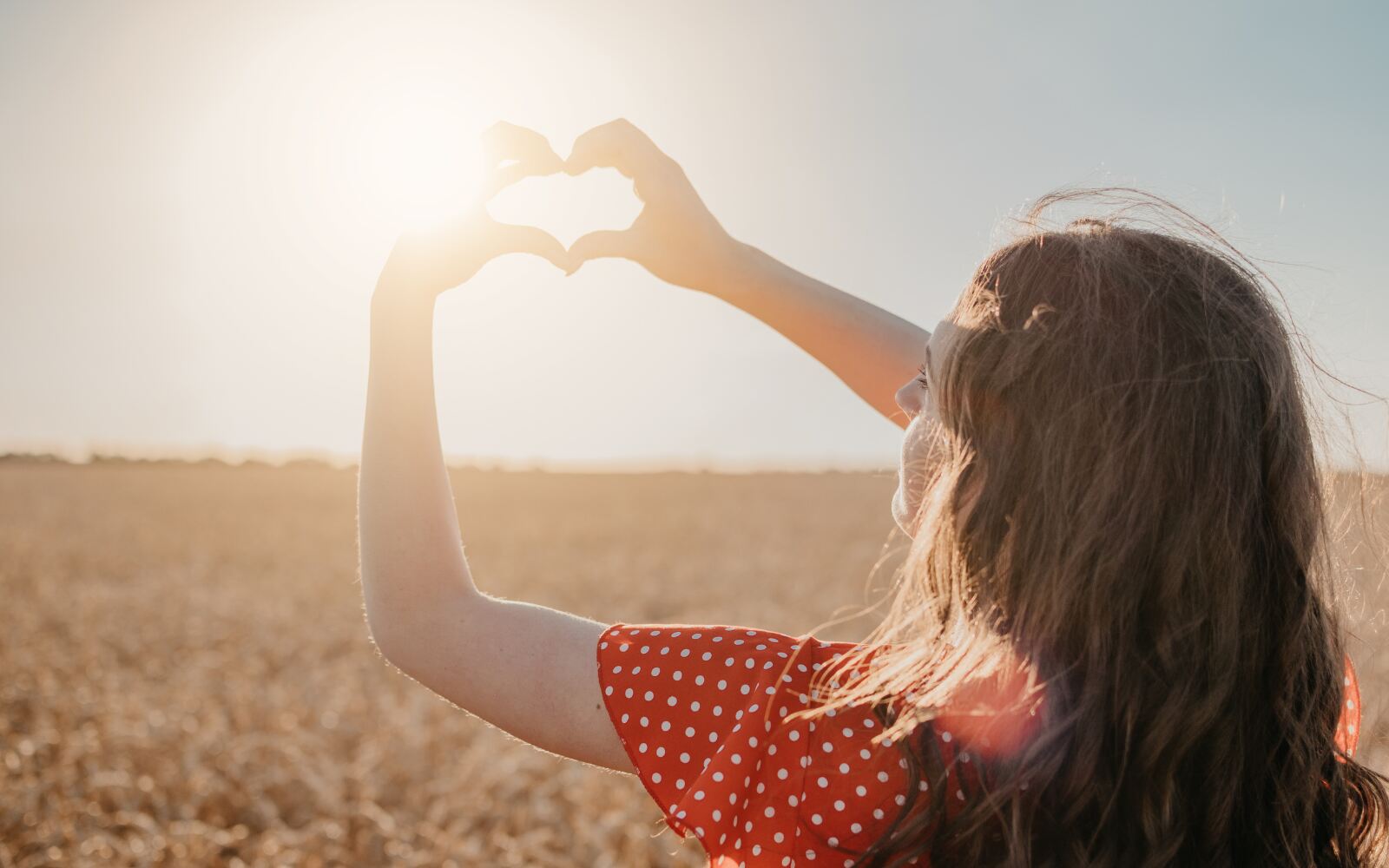 a woman is outside holding her hands up towards the sun, making a heart - she's wearing a red polka dot dress and her hair is flowing in the wind