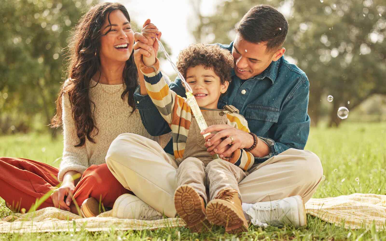 a mom, dad, and their son are sitting outside in the sunshine, the son is on the dad's lap pulling a tube of bubbles out of its container