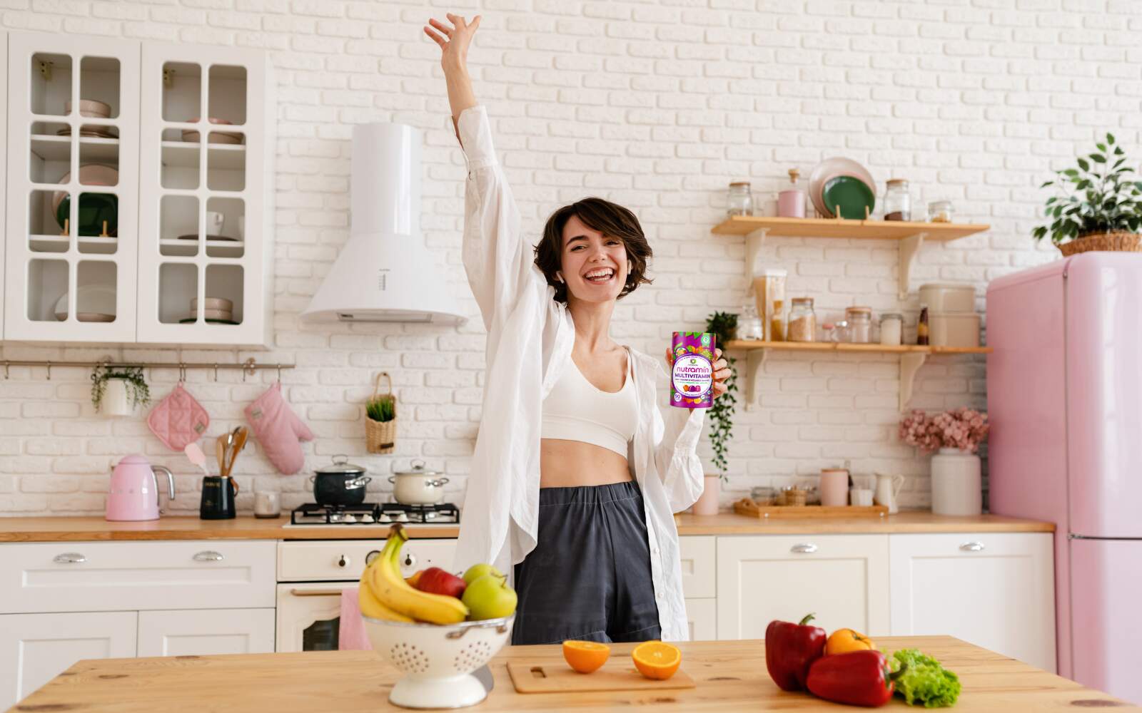 a healthy woman is standing in a brightly lit kitchen, one arm is extended to the roof celebrating while her other hand holds a bottle of sugar free nutramin vitamin gummies